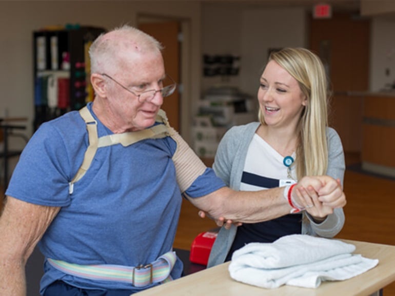 An older man with a supportive harness around his shoulders is performing a rehabilitation exercise at a wooden table with assistance from a smiling female therapist.