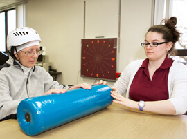 Female patient wearing safety helmet holding a large blue foam roller on a therapy table.