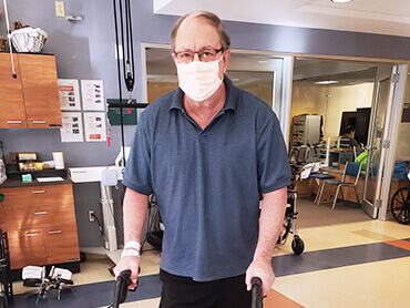 Robert using a rolling walker and standing in a therapy gym.
