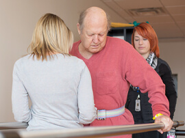 Two female therapists help steady male patient using parallel bars to walk.