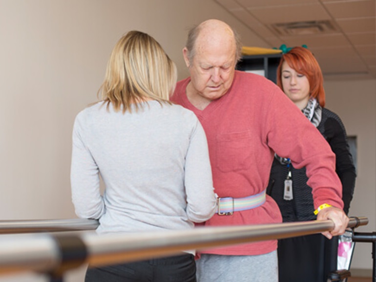 Two female therapists spot a male patient using parallel bars to balance while walking.