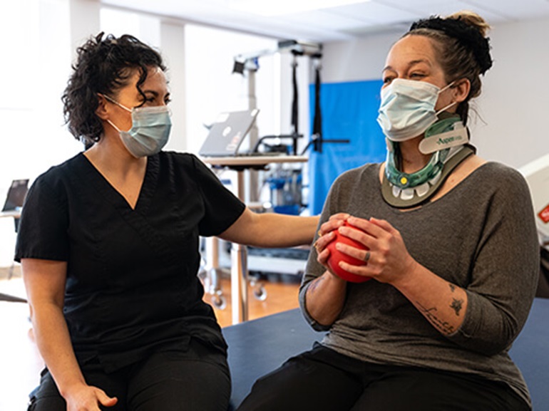 Female patient wearing a neck brace and Covid mask while holding a small red ball in her hands.