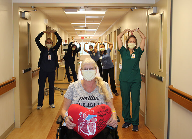 Debra sitting in a wheelchair holding a heart-shaped pillow with nurses in background make heart shapes with their arms.